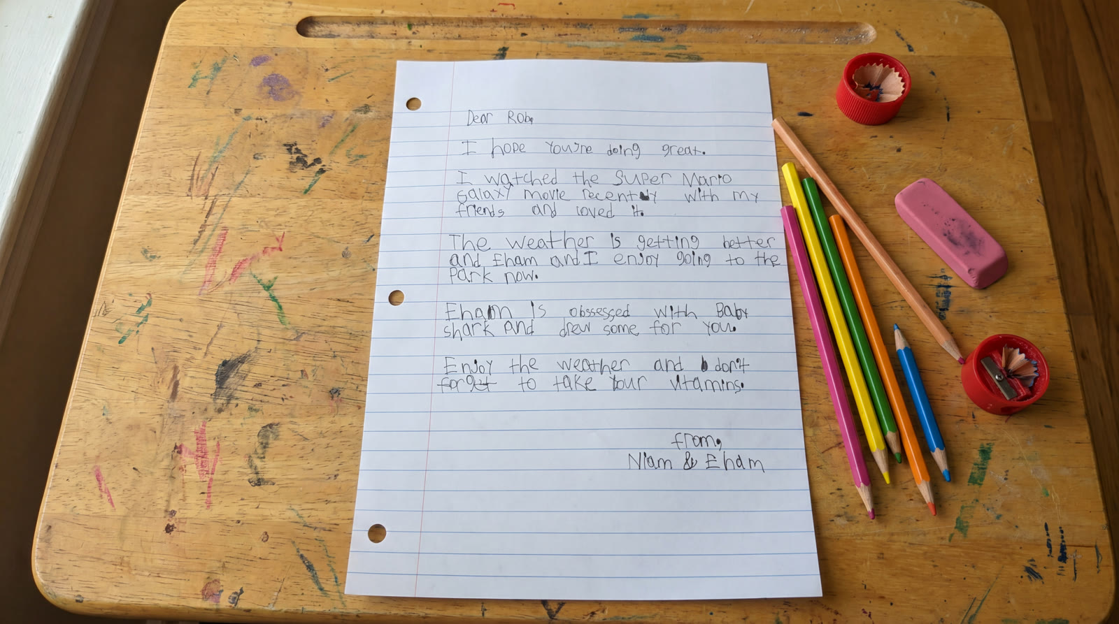 A handwritten letter from Niam, age 7, on lined paper, resting on a wooden lap-desk with colored pencils and erasers